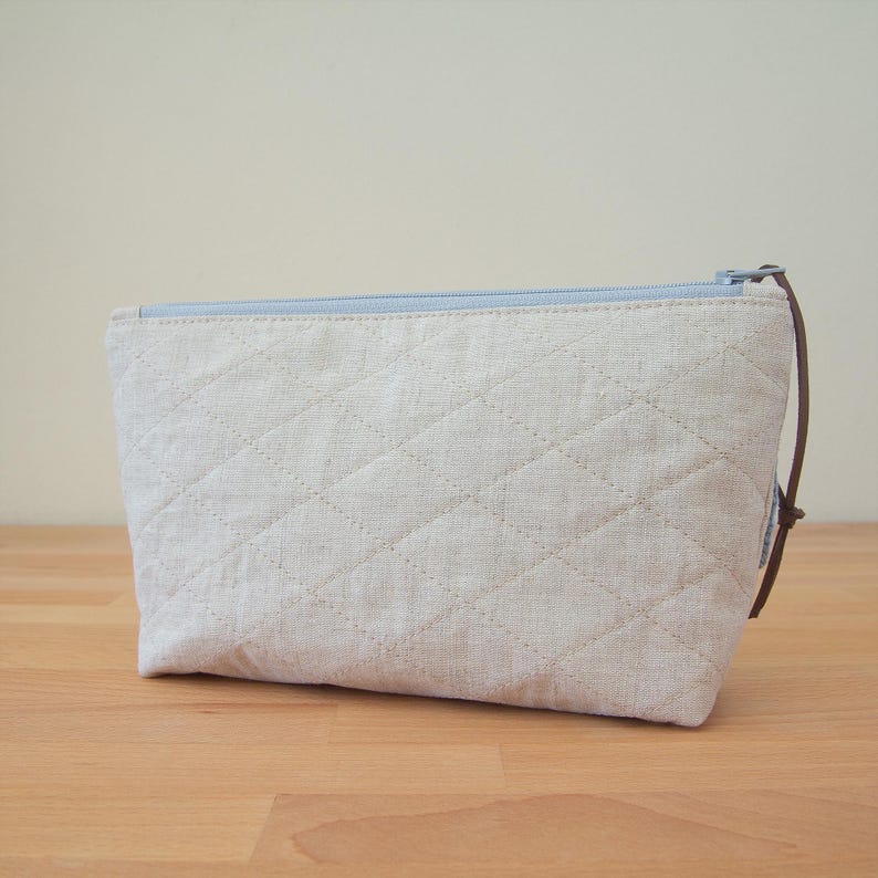 May include: A light blue zippered pouch with a quilted linen exterior. The pouch is made of a natural fabric and has a brown leather pull tab.