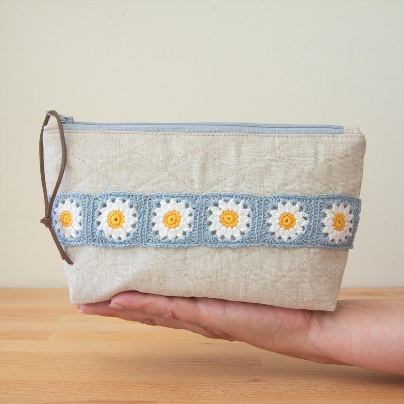 May include: A light blue and white crocheted daisy design adorns a beige linen zippered pouch. The pouch is quilted and has a brown leather zipper pull.