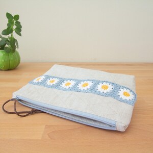 May include: A light blue zippered pouch with a crocheted daisy trim. The pouch is made of a light beige linen fabric.