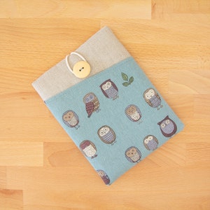 Op de afbeelding: Een blauwe stoffen hoes met een beige flap en een knoopsluiting. De hoes is versierd met een patroon van uilen in verschillende kleuren en ontwerpen.