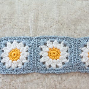 May include: A close-up of a crocheted border with blue and white daisy flowers. The flowers have yellow centers and are arranged in a repeating pattern.