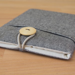May include: A gray and white fabric tablet sleeve with a wooden button closure. The sleeve is folded closed and the button is fastened.