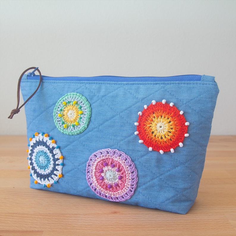 May include: A blue denim zippered pouch with four colourful crocheted circles attached. The circles are in shades of green, orange, purple, and blue.