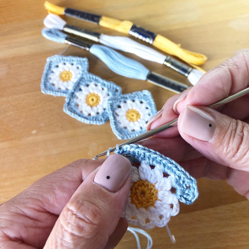 May include: Close-up of a person crocheting a blue and white daisy granny square with a crochet hook. The person is holding the crochet hook in their right hand and the yarn in their left hand. The crochet hook is resting on the edge of the granny square. The yarn is a light blue color.