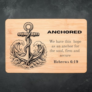 Puede incluir: Tabla de cortar rectangular de madera con un diseño de ancla grabado con láser y la palabra "ANCHORED". El ancla está rodeada de olas. El texto de abajo dice "Tenemos esta esperanza como ancla del alma, firme y segura Hebreos 6:19".