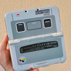 Może przedstawiać: Jasnoniebieska konsola do gier przenośna Nintendo 3DS XL z motywem Super Nintendo. Konsola posiada logo Super Nintendo, przyciski zasilania i resetowania oraz napis "EJECT".