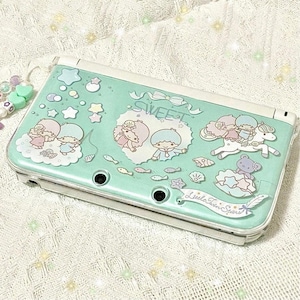 Puede incluir: Una consola Nintendo 3DS verde menta con una funda protectora transparente. La funda está decorada con ilustraciones de los personajes Little Twin Stars, estrellas, nubes y peces. La palabra "SWEET" está escrita en la parte superior.