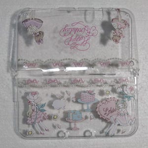 May include: Clear plastic case with a hinged lid, decorated with pink and white illustrations. The design includes a cake, a bunny, and a girl in a dress. The words "Happy Birthday" are written in pink script. The word "Lolita" is also visible.