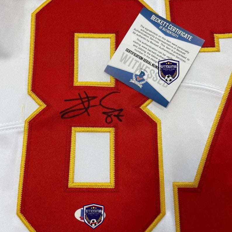 May include: A white sports jersey featuring a large red number eight with yellow trim. The number is signed in black ink. A Beckett Certificate of Authenticity card is placed on top, and a small shield-shaped logo is visible at the bottom of the number.