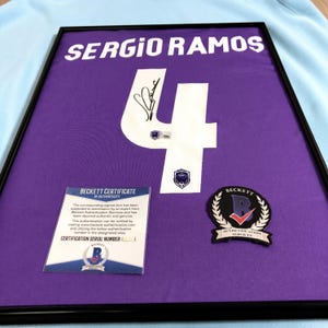 Może przedstawiać: Oprawiona fioletowa koszulka sportowa z nazwiskiem SERGIO RAMOS i numerem 4 w kolorze białym. Numer 4 zawiera czarny autograf, naklejkę uwierzytelniającą Beckett oraz mały emblemat. Dołączony jest certyfikat autentyczności Beckett.