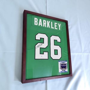 Może przedstawiać: Oprawiona w ramę zielona koszulka sportowa z nazwiskiem BARKLEY i numerem 26 w kolorze białym z czarnymi obwódkami. Koszulka zawiera czarny podpis na numerze 26, małą naklejkę autentyczności oraz prostokątną etykietę z certyfikatem.