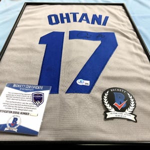 Pode incluir: Uma camisola desportiva cinzenta emoldurada com o nome OHTANI e o número 17 em azul. A camisola inclui um autógrafo a tinta preta, um autocolante de autenticação Beckett no número, um certificado de autenticidade e um emblema Beckett.