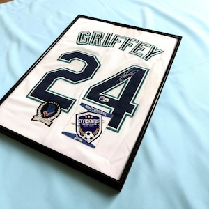 May include: A framed white sports jersey featuring the name GRIFFEY and the number 24 in navy blue with teal trim. The jersey includes a signature on the number 24, a Beckett Authentication Services patch, and a Beckett Certificate of Authenticity.
