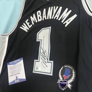 May include: A black basketball jersey featuring the name WEMBANYAMA and the number 1 in white with silver trim. The number 1 includes a black ink signature and a small authentication sticker. A Beckett Certificate of Authenticity is included.