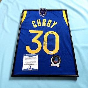 May include: A framed blue and yellow basketball jersey with the name CURRY and number 30 printed on the back. The number 30 features a black ink autograph and the handwritten text GOAT. Includes a Beckett Certificate of Authenticity and logo.