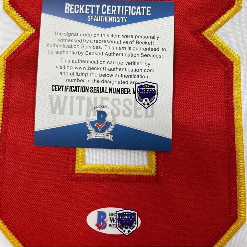 May include: A Beckett Certificate of Authenticity card with white and blue text placed over a red fabric patch with a yellow border. The card features the Beckett logo, a soccer ball emblem, and the text Certification Serial Number.