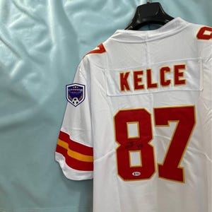 May include: A white sports jersey featuring the name KELCE and the number 87 in red with yellow trim on the back. The jersey includes a signature on the number 87, a small authentication sticker, and a patch on the sleeve with the text EFFICIENTUS.