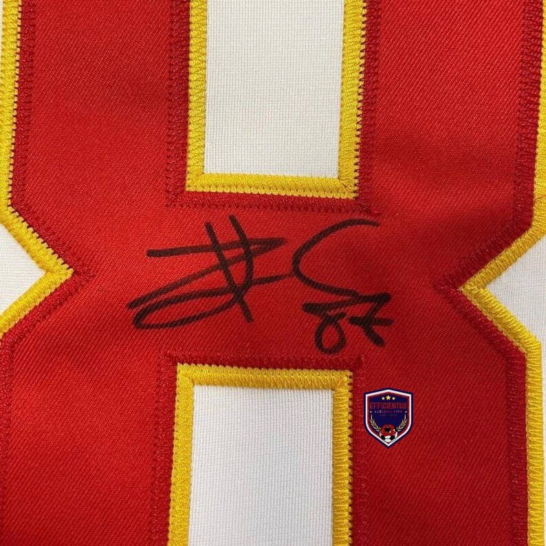 May include: A red fabric patch featuring a large white number eight with a thick yellow embroidered border. A black ink signature is written across the center of the number. A small shield-shaped logo with the text EFFICIENTES is visible.