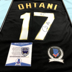 May include: A black jersey framed in a dark border, featuring the name OHTANI and the number 17 in gold and white lettering. A signature is visible on the number 17, accompanied by a Beckett Certificate of Authenticity and a Beckett authentication sticker.