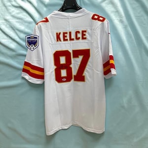 May include: A white sports jersey featuring the name KELCE and the number 87 in red with gold outlines on the back. The sleeves have red and gold stripes. A small logo patch with the text EFFICIENTUS is on the left shoulder area.