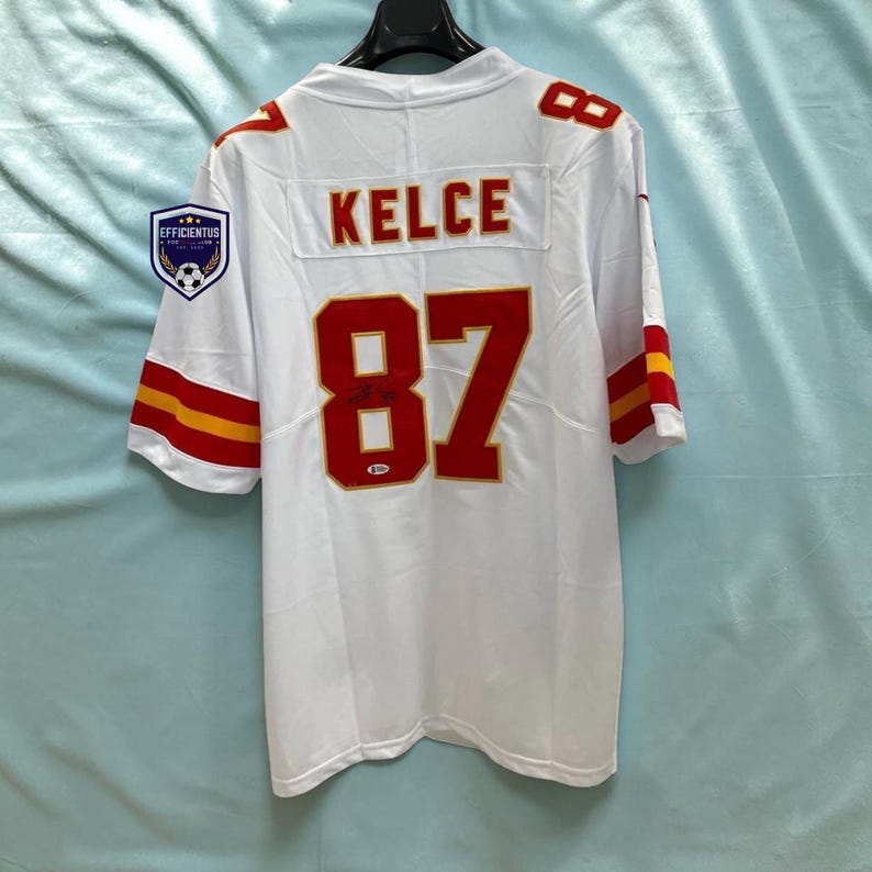 May include: A white sports jersey featuring the name KELCE and the number 87 in red with gold outlines on the back. The sleeves have red and gold stripes. A small logo patch with the text EFFICIENTUS is on the left shoulder area.