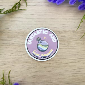 Könnte beinhalten: Ein runder Aufkleber mit einem violetten Hintergrund und weißem Text, der "Fantastisch und Nicht-Binär" lautet. Eine kleine Flasche mit einer gelben, weißen und violetten Flüssigkeit befindet sich im Kreis. Die Flasche hat einen Korkverschluss und einen Stern darüber.