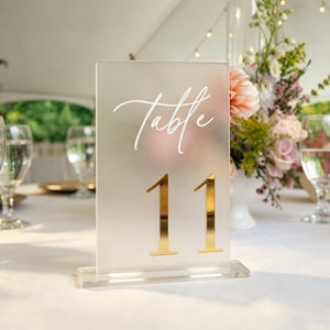May include: A rectangular frosted acrylic table sign featuring the white cursive text "Table" above the large, reflective gold-colored number "11". The sign is supported by a clear acrylic base and stands upright on a white tablecloth.
