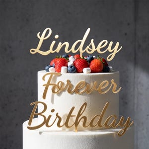 Puede incluir: Un adorno para pastel de color dorado con el nombre Lindsey y la frase Forever Birthday en letra cursiva. El adorno está colocado sobre un pastel con glaseado blanco decorado con fresas rojas frescas, arándanos y malvaviscos.