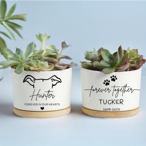 May include: Two white ceramic planters with succulents, each featuring a wooden base. One planter has a dog silhouette and the name "Hunter" with the text "Forever in our hearts." The other says "forever together" with paw prints and the name "Tucker."