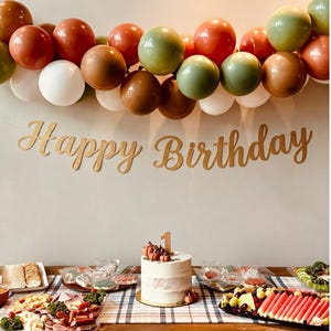 Könnte beinhalten: Ein Party-Deko-Set mit einem goldglitzernden "Happy Birthday"-Banner und einer Ballongirlande in Salbeigrün, Terrakotta, Braun und Weiß. Ein weißer Kuchen mit einer "1" als Tortenstecker steht auf einem Tisch mit verschiedenen Speisen.