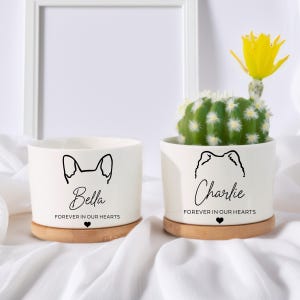 Può includere: Due vasi in ceramica bianca con sottovasi in bambù. Un vaso presenta un contorno di orecchio di cane e il nome "Bella" con "Forever in our hearts" sotto. L'altro ha un contorno di cane e il nome "Charlie".
