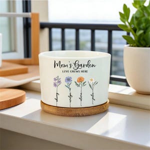 May include: A white ceramic Mom's Garden planter with a bamboo base. The planter features the text "Mom's Garden LOVE GROWS HERE" and illustrations of four flowers with names below them. A small white planter with a green plant is visible in the background.