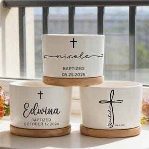 Puede incluir: Tres macetas de cerámica blanca con bases de madera. Cada maceta presenta una cruz negra y texto personalizado, incluyendo nombres y fechas de bautizo. Las macetas son cilíndricas y se asientan sobre platillos redondos de madera.
