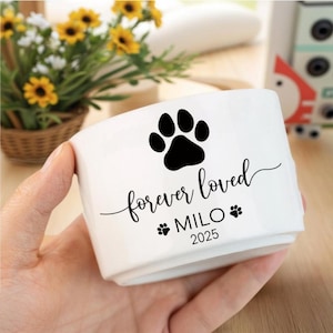 May include: A white ceramic pet bowl with a black paw print and the words "forever loved" in cursive. The bowl also includes the name "MILO" and the year "2025". The bowl is held in a person's hand.