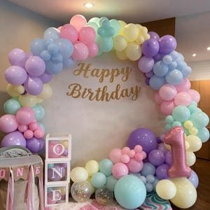 May include: A large balloon arch in pastel shades of pink, blue, purple, and yellow surrounds a gold glittery "Happy Birthday" sign. Three stacked clear boxes spell "ONE" with pink balloons inside, and a large metallic pink "1" balloon.