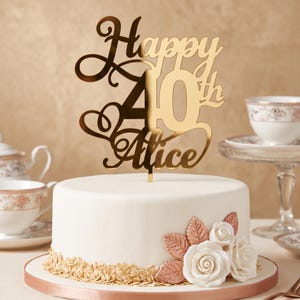Peut inclure: Une décoration de gâteau en acrylique doré portant l'inscription Happy 40th Alice dans une police cursive. La décoration est placée sur un gâteau rond blanc orné de roses en glaçage blanc et de feuilles couleur cuivre métallique à la base.