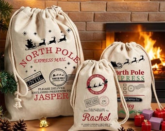 May include: Three beige canvas drawstring gift bags with North Pole Express Mail graphics. The bags feature red and black text, including the names Jasper and Rachel, reindeer silhouettes, and postal-style stamps with holiday dates.