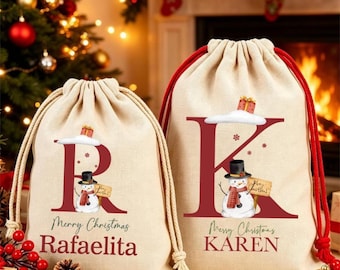 Puede incluir: Dos bolsas de regalo de tela de color beige con cordón. La bolsa más pequeña tiene una letra R roja grande, un muñeco de nieve y el nombre RAFAELITA. La bolsa más grande tiene una letra K roja grande, un muñeco de nieve y el nombre KAREN. Texto Merry Christmas.