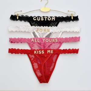 May include: Four pairs of thong underwear in black, white, pink, and red, each with a different gold text design: "CUSTOM", "JOHN", "ALL YOURS", and "KISS ME". The underwear are displayed on a gold hanger.