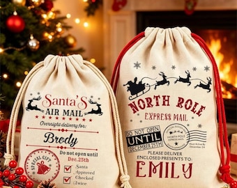 May include: Two beige drawstring canvas gift bags with red cords. One bag features Santa's Air Mail text with the name Brody, while the other displays North Pole Express Mail text with the name Emily, both decorated with reindeer and holiday graphics.