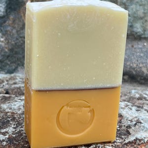 Pode incluir: Uma barra de sabão de duas cores. A metade superior é creme esbranquiçado e a metade inferior é amarelo dourado. A metade inferior tem um logotipo circular em relevo com a palavra "Farmhouse". O sabão é retangular e parece ser feito à mão.