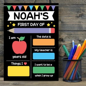 May include: A black chalkboard sign titled Noah's First Day Of with colourful bunting, a pencil graphic, and an apple icon. It features blank fields for age, date, teacher, interests, and future career goals, accompanied by a black wire pencil cup.