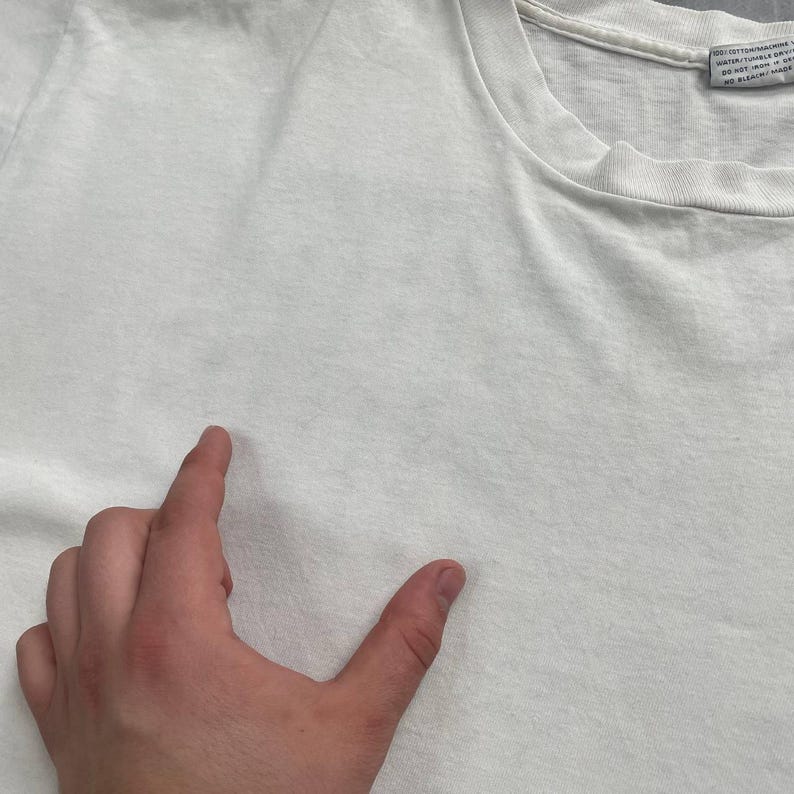 May include: A close-up of a white t-shirt. The shirt is made of a soft material and has a crew neck. A hand is pointing at the shirt.