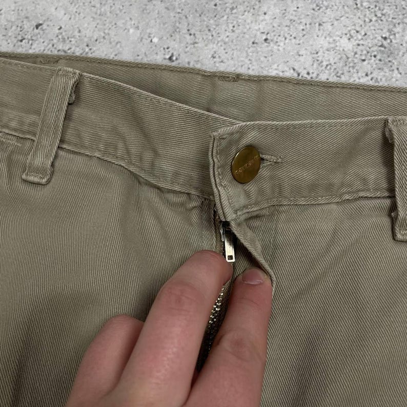 May include: Close-up of khaki-colored pants with a button and zipper closure. The pants have belt loops and a metal zipper. The button has the brand name engraved on it. The fabric appears to be a durable cotton blend.