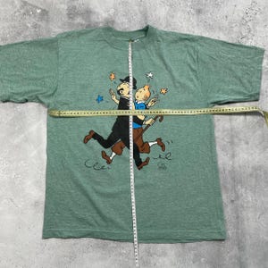 Pu&ograve; includere: T-shirt verde con una grafica a cartoni animati di due personaggi che si abbracciano, con stelle e altre piccole illustrazioni. I personaggi sono in nero, marrone e blu. La t-shirt &egrave; misurata con un metro a nastro.