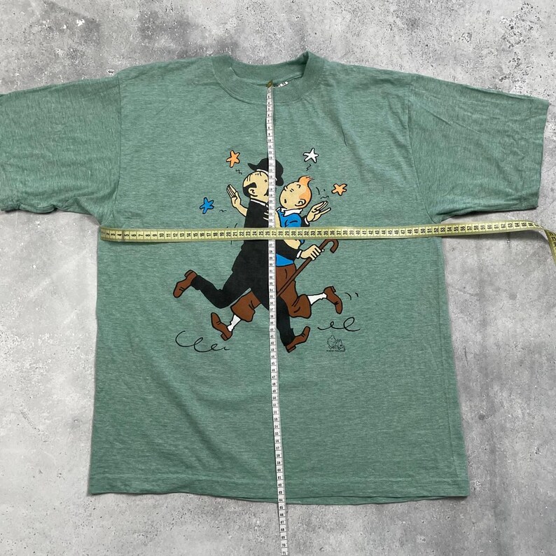 Pu&ograve; includere: T-shirt verde con una grafica a cartoni animati di due personaggi che si abbracciano, con stelle e altre piccole illustrazioni. I personaggi sono in nero, marrone e blu. La t-shirt &egrave; misurata con un metro a nastro.