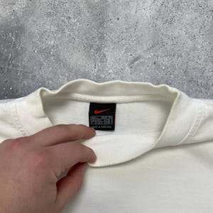 Peut inclure: Gros plan sur un col de t-shirt blanc avec une &eacute;tiquette noire portant un logo Nike rouge. L'&eacute;tiquette comprend des informations de taille et le texte "MADE IN PORTUGAL". Le t-shirt est sur fond gris.