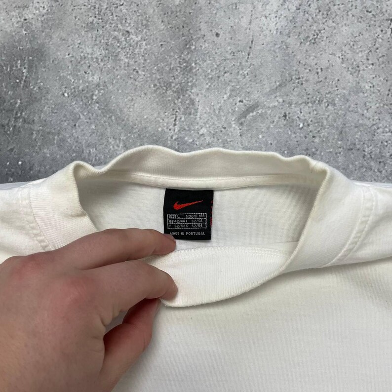 Peut inclure: Gros plan sur un col de t-shirt blanc avec une &eacute;tiquette noire portant un logo Nike rouge. L'&eacute;tiquette comprend des informations de taille et le texte "MADE IN PORTUGAL". Le t-shirt est sur fond gris.