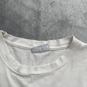 May include: Close-up of a white t-shirt collar with a tag. The tag reads "100% COTTON/MACHINE WASH/COLD WATER/TUMBLE DRY/LOW HEAT/DO NOT IRON IF DECORATED/NO BLEACH/MADE IN USA."