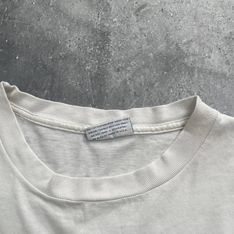 May include: Close-up of a white t-shirt collar with a tag. The tag reads "100% COTTON/MACHINE WASH/COLD WATER/TUMBLE DRY/LOW HEAT/DO NOT IRON IF DECORATED/NO BLEACH/MADE IN USA."